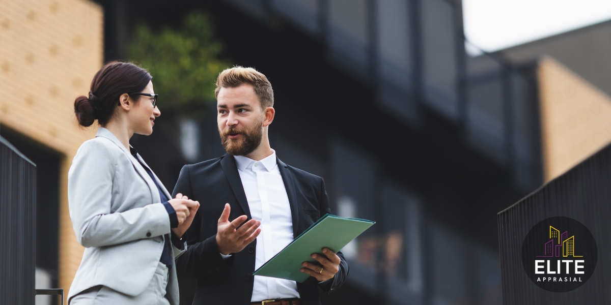 Two business professionals—a woman in a light gray blazer and a man in a dark suit—stand outside a modern building discussing paperwork, with the man holding a green folder. The Elite Appraisal logo (stylized purple and yellow city skyline with the words “ELITE APPRAISAL”) appears in the bottom right corner.