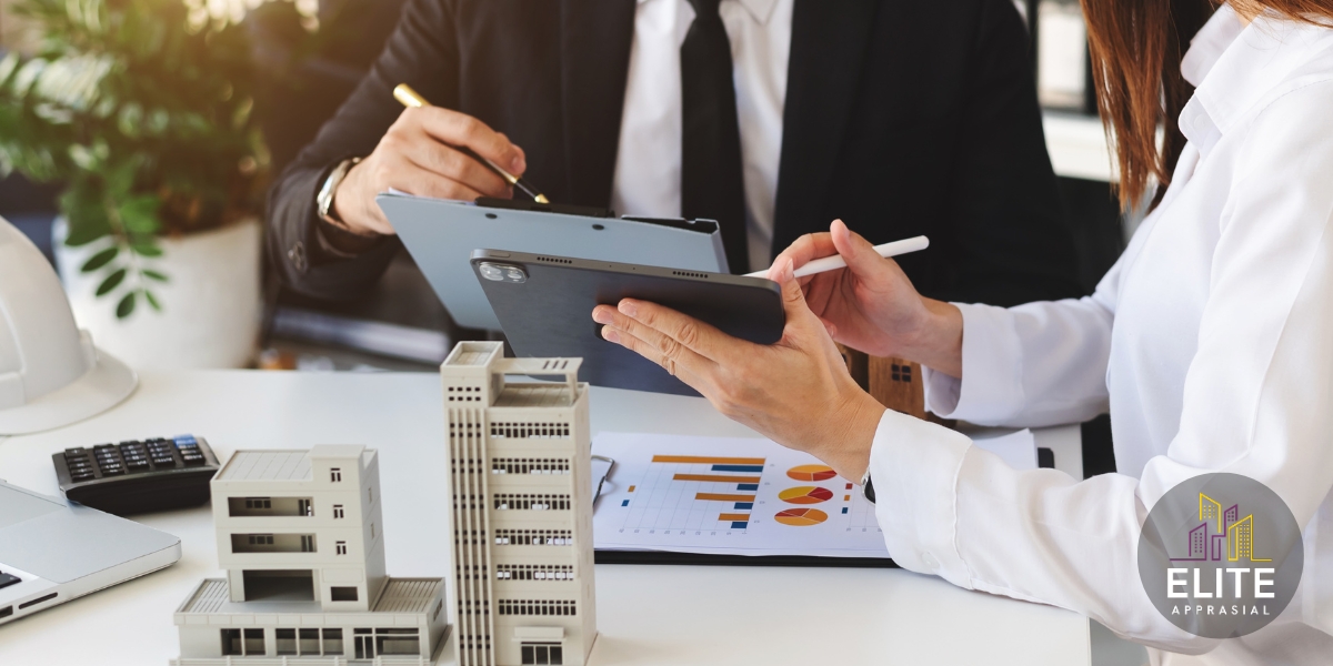 A close-up of two professionals—one in a dark suit holding a clipboard and pen, the other in a white shirt using a stylus on a tablet—discussing data at a desk. On the desk are scale models of multi-story buildings, a calculator, a laptop, and a printed chart with bar and pie graphs. The Elite Appraisal logo (stylized purple and yellow skyline with “ELITE APPRAISAL”) appears in the lower right corner.