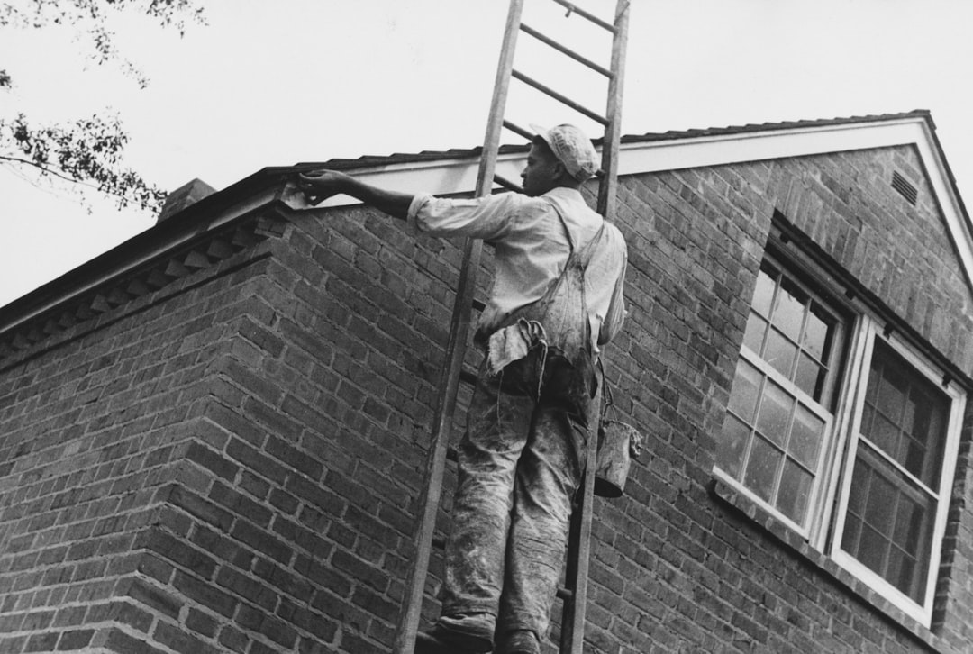 A roofing contractor stands on a residential roof, holding a clipboard and pen while carefully inspecting the shingles for storm damage.